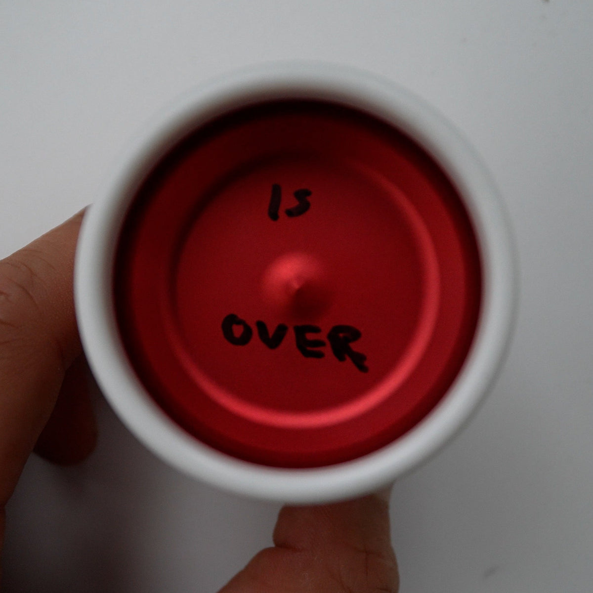 A hand holding a white cup from above, showing a bright red interior with the handwritten words 'IS OVER' in black marker on a plain light background.
