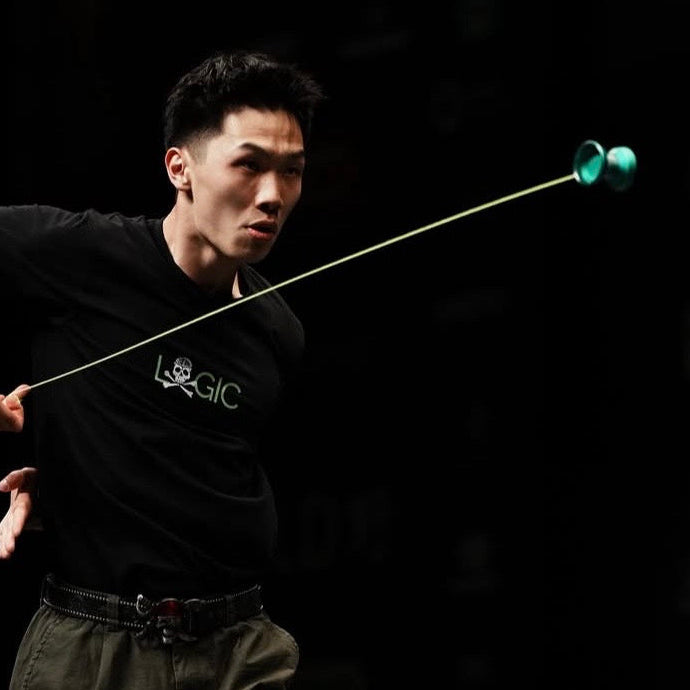 A man performing with a long, thin yo-yo on a dark stage, wearing a black t-shirt and olive green cargo pants.